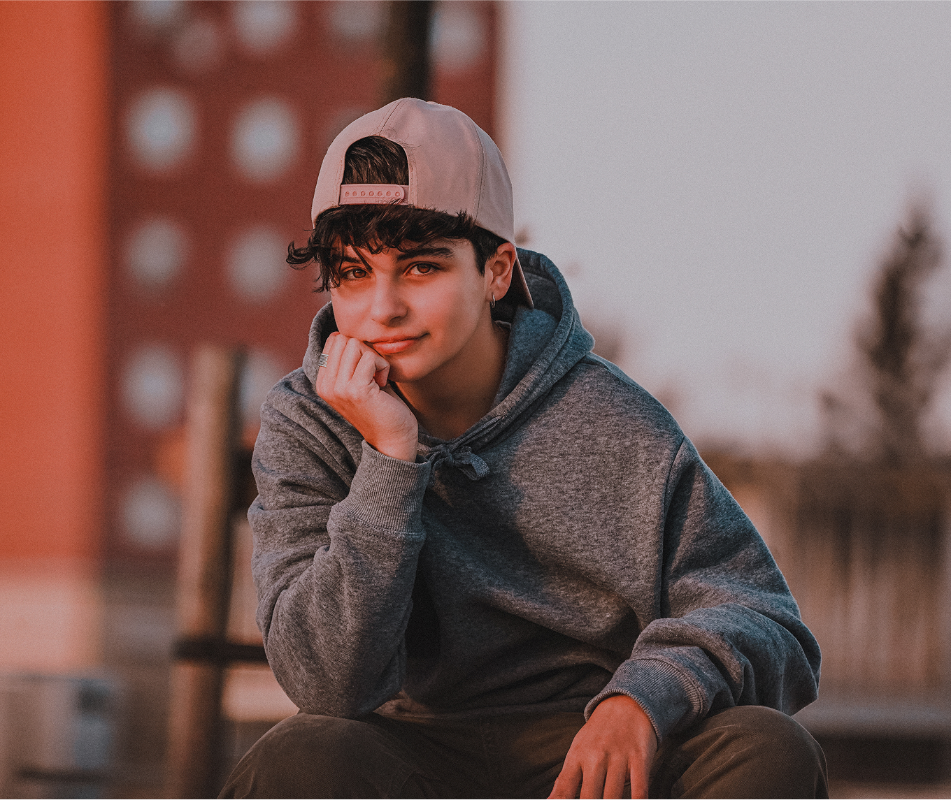 Eine Person mit grauem Kapuzenpulli und umgekehrter Mütze sitzt im Freien, das Kinn auf die Hand gestützt, mit einem verschwommenen Gebäude und Bäumen im Hintergrund.