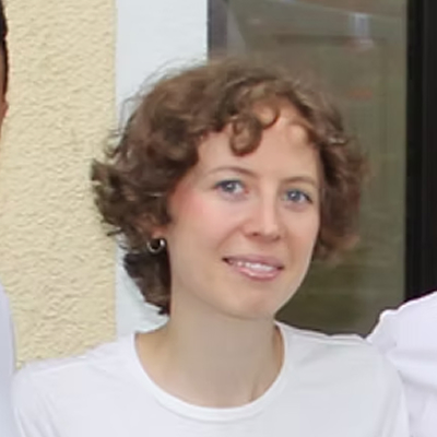 Eine Person mit kurzen lockigen braunen Haaren und blauen Augen, die ein weißes Hemd und kleine Ohrringe trägt, steht vor einer beigen Wand und einem Fenster.