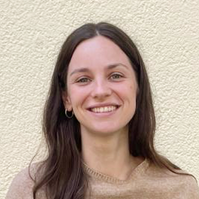 Eine Person mit langen braunen Haaren, die einen beigen Pullover und kleine Ohrringe trägt, lächelt vor einer hellen, strukturierten Wand.