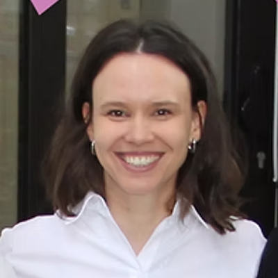 Eine Person mit schulterlangen braunen Haaren und Ohrringen lächelt, trägt ein weißes Hemd mit Kragen und steht in einem Innenraum vor einer Glastür.