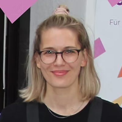 Eine Person mit schulterlangem blondem Haar, Brille und Haarknotenfrisur lächelt und blickt in die Kamera vor einem Hintergrund mit geometrischen Formen.