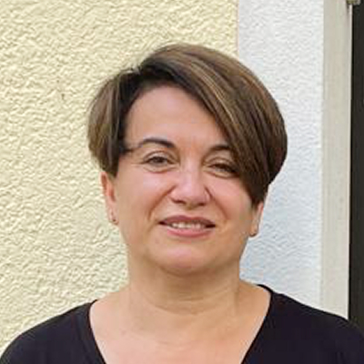 Eine Person mit kurzen braunen Haaren steht vor einer hellen, strukturierten Wand, trägt ein schwarzes Oberteil und schaut in die Kamera.